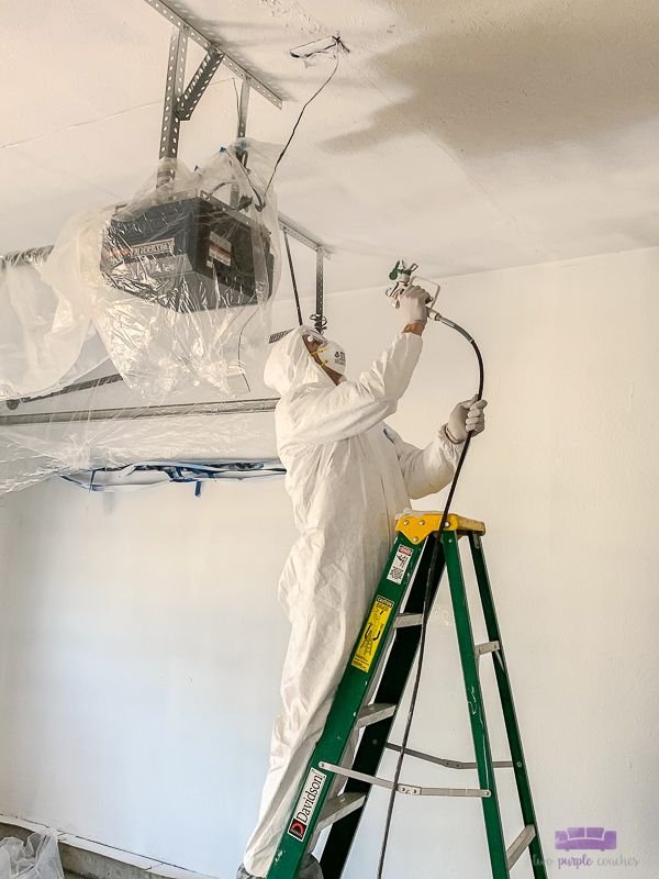 Pintor con equipo de protección aplica pintura al techo de un garaje con un pulverizador.