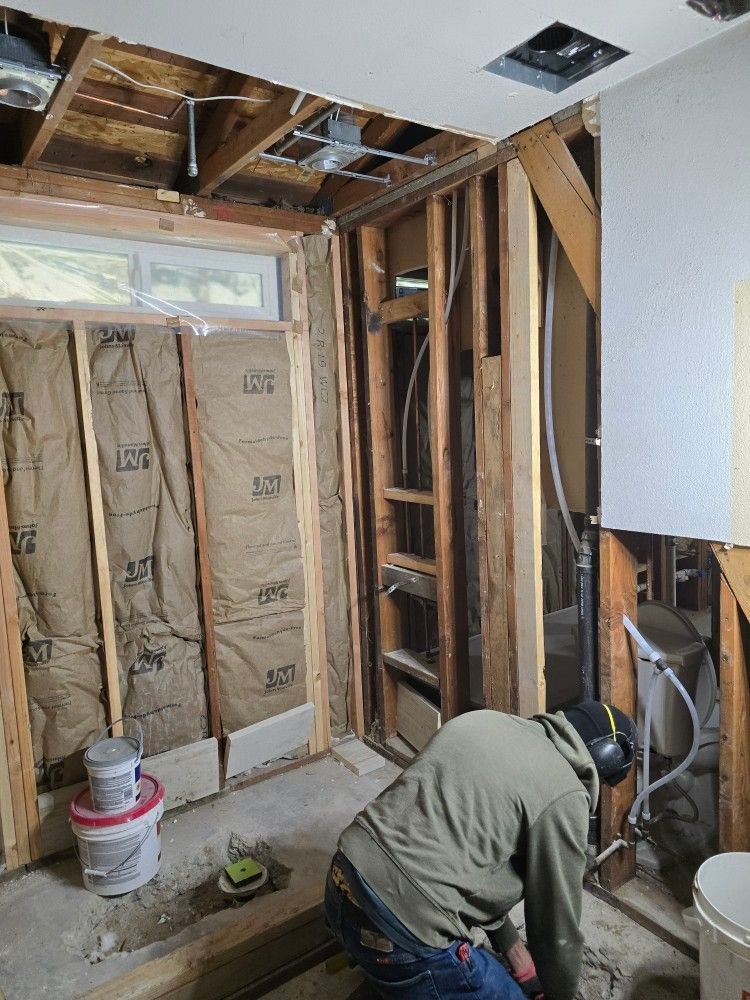 Trabajador realiza labores de plomería en una habitación con tuberías y estructura de madera expuestas.