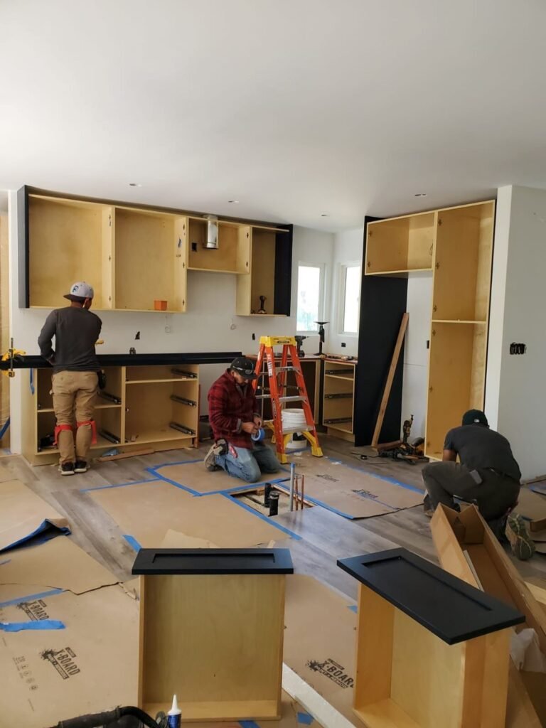Tres trabajadores instalan gabinetes de madera y frentes oscuros durante la remodelación de una cocina.